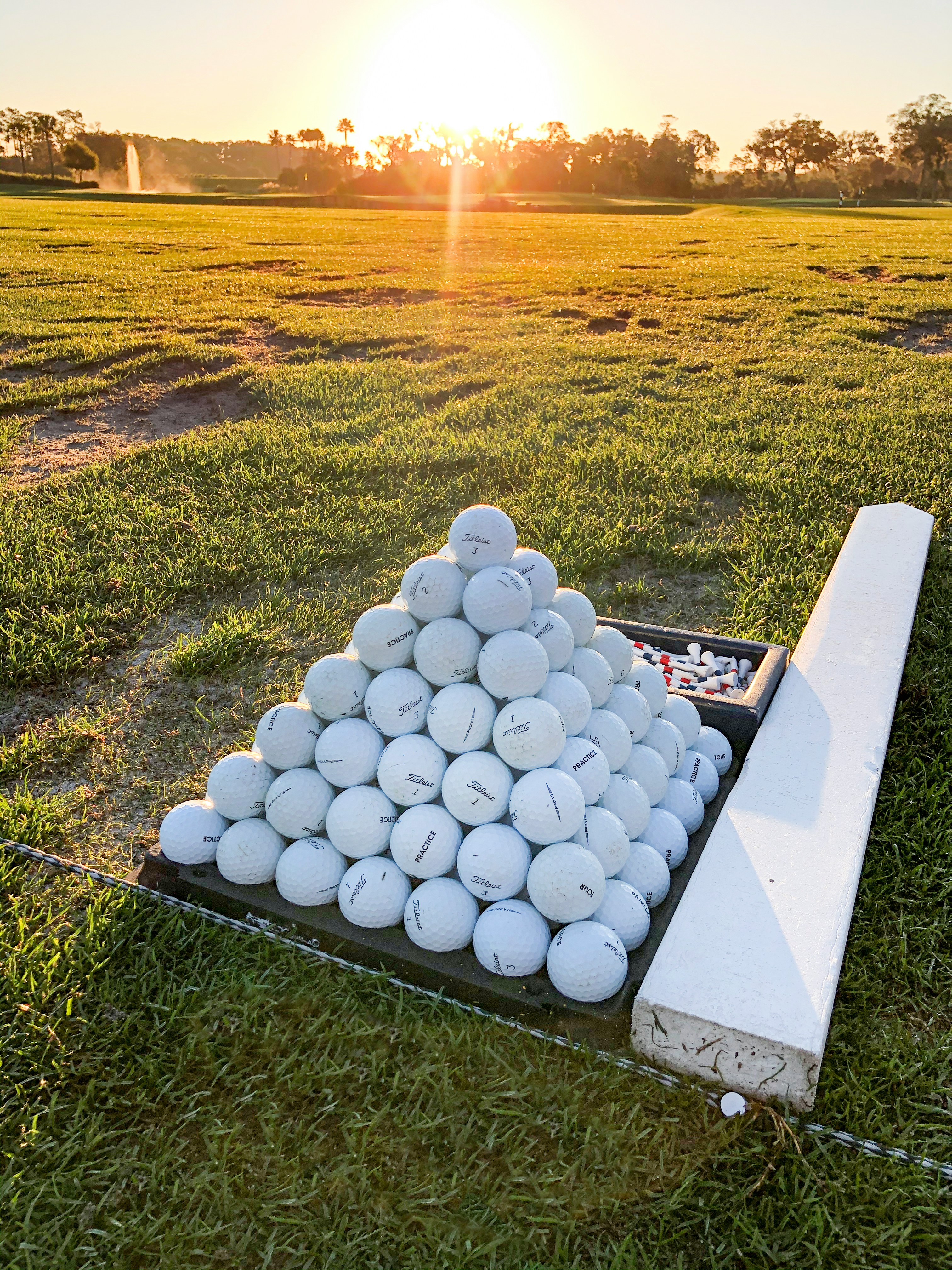 A pyramid of practice golf balls on a driving range mat at sunset, with lens flare and tree silhouettes on the horizon